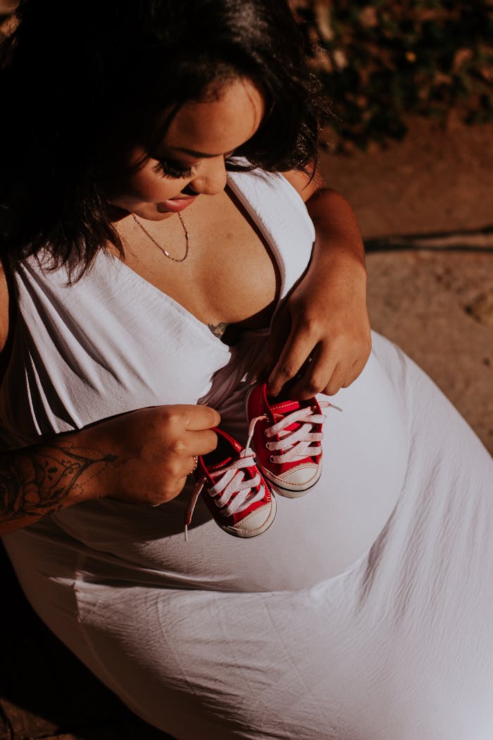 A pregnant woman in a white dress holding red baby sneakers, symbolizing anticipation and motherhood.