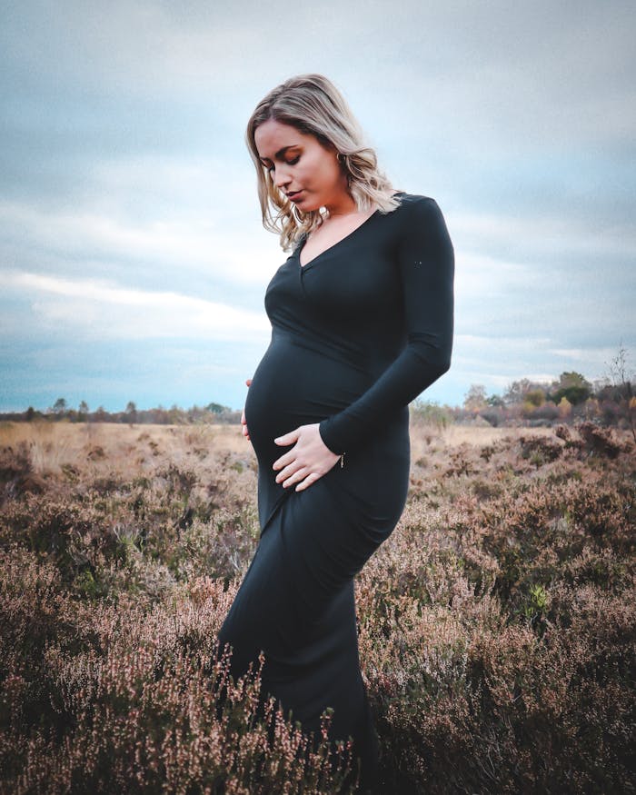 our-services-3 Pregnant woman in black dress posing outdoors in a tranquil field setting.