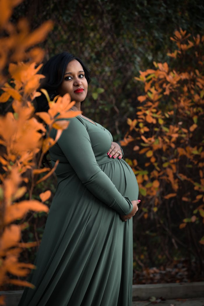 our-services-2 Expectant mother in a green dress posing elegantly among autumn leaves in a serene outdoor setting.