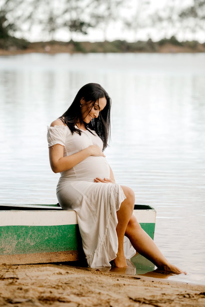 why-choose-us Expectant mother in a white dress sits by the beach, embracing maternity.