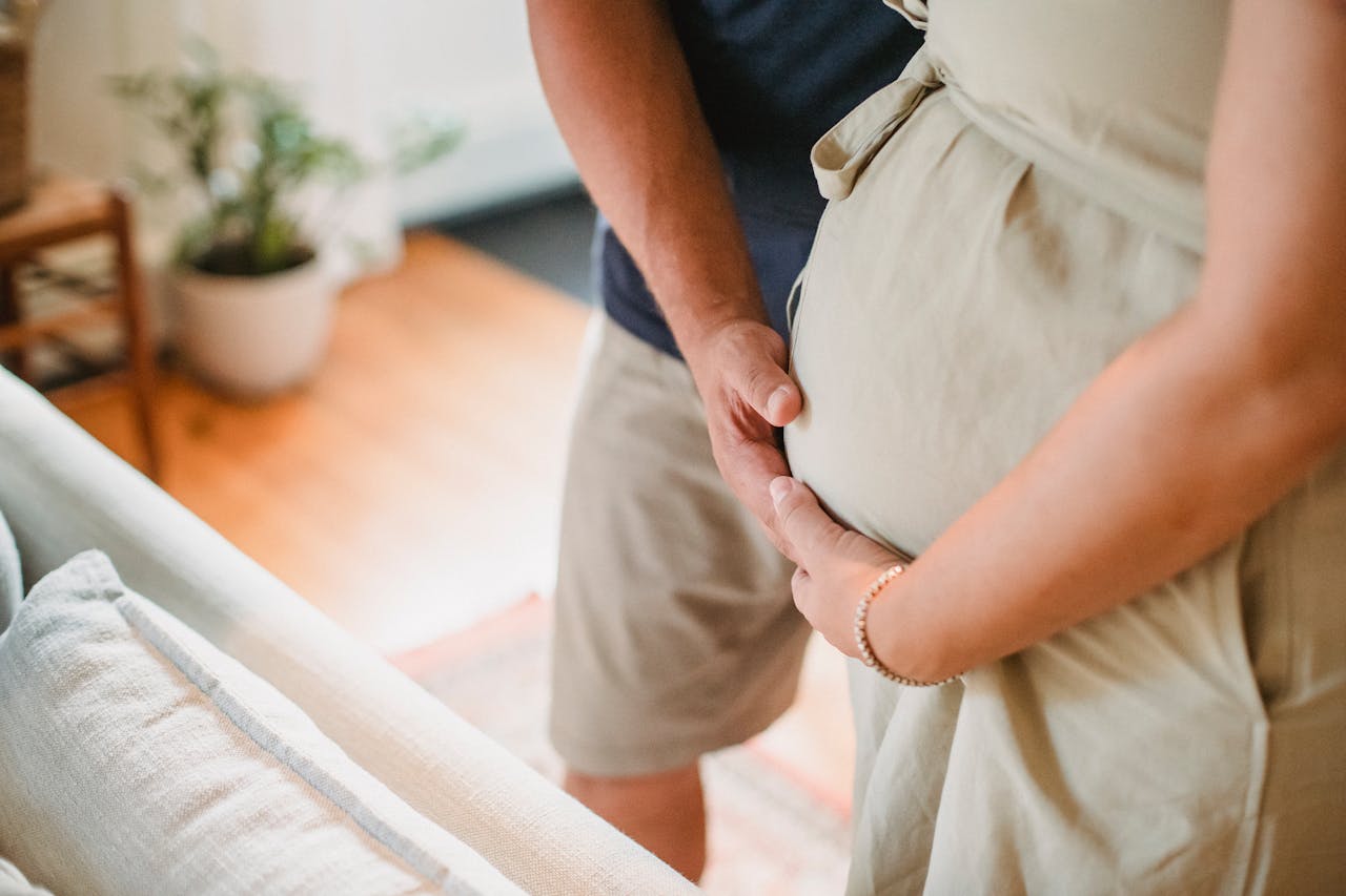gallery-6 From above of crop faceless couple in casual outfit standing in light living room and touching pregnant belly gently