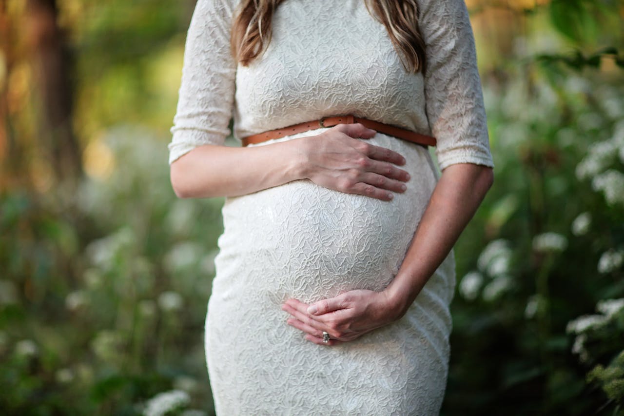 hero-img-02 A pregnant woman in a lace dress gently cradling her belly in an outdoor setting.